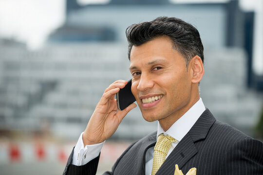 Smiling smartly dressed Asian / Middle eastern businessman holding a mobile phone to his ear, in an outdoor city location.