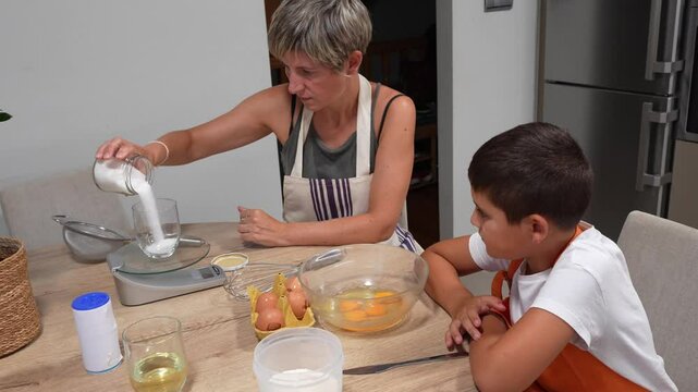 Caring mother guiding son while precisely measuring sugar on kitchen scale, sharing culinary skills during sponge cake preparation