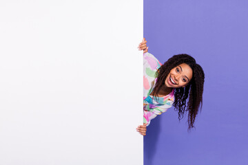 Smiling young woman with curly hair playfully peeking around a white panel against a vibrant purple...