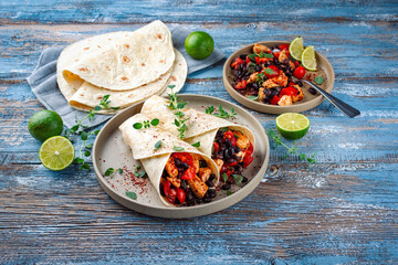 Mexican style burritos - tortilla wrap with spicy chicken meat breast filet nuggets, vegetables and black olives served as close-up with hot spices on a design plate