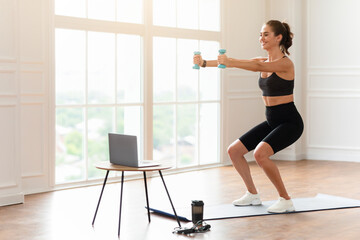 Obraz premium Positive fit lady exercising and squatting with two dumbbells in front of pc on coffee table at home, smiling fitness blogger recording video for her followers having personal virtual training