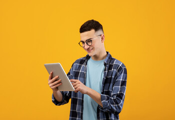 Online work, communiction, education. Cool young man browsing web on tablet pc over orange studio background. Funky student in glasses using touch pad, watching videos, posting in social media