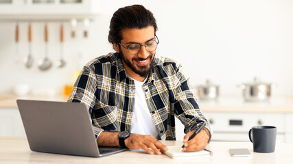 Positive arab guy with glasses looking for job online, sitting at table, using laptop and taking notes, kitchen interior, copy space. Excited indian man student working online from home, remote job