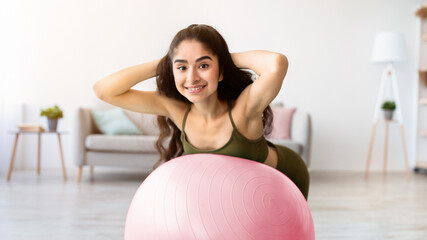 Happy young Indian woman doing exercises with fitness ball, strengthening her back muscles at home. Millennial Asian lady in sportswear having domestic training during covid quarantine