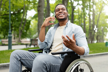 Joyful handicapped black man in wheelchair talking on smartphone at green park. Portrait of...