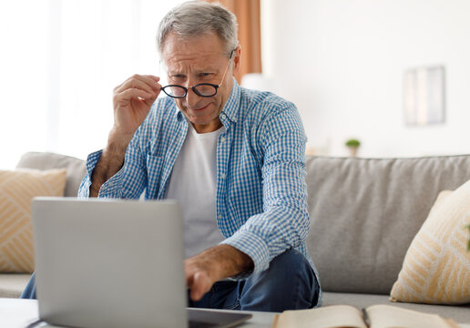 Poor Eyesight. Senior Man Squinting Eyes Using Laptop Wearing Eyeglasses Having Problems With Vision Sitting On Couch. Ophtalmic Issue, Bad Sight In Older Age, Macular Degeneration Concept
