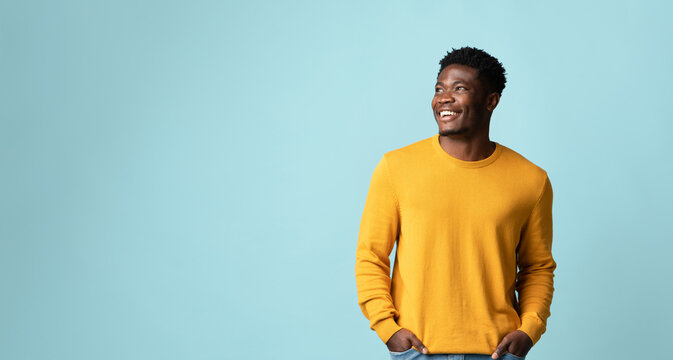 Happy millennial black man in casual outfit holding hands in pockets, looking at copy space for text or advertisement and smiling, recommending nice offer or discounts, studio shot on blue, panorama