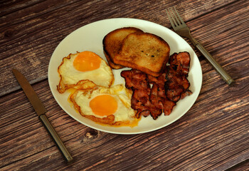 Fried eggs with bacon and toast on a dark wooden table, cutlery nearby.