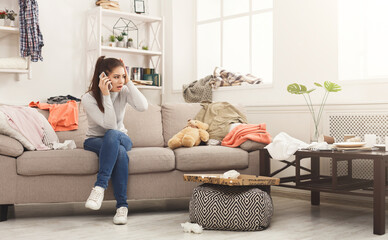 Desperate helpless woman sitting on sofa in messy living room. and talking on mobile, surrounded by many stack of clothes. Disorder and mess at home, copy space