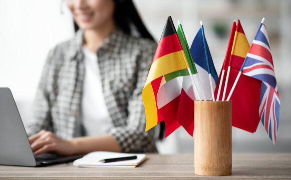 Cropped of asian young woman foreign language teacher sitting at workdesk and using laptop, selective focus on international flags. Foreign language online course, class, e-education concept