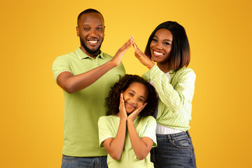 Family care, protection and insurance concept. Young black parents making symbolic roof of hands above little daughter while standing over yellow background, studio shot © Prostock-studio