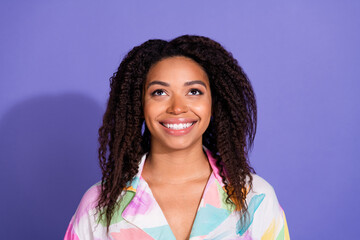 Charming young woman with curly hair in colorful outfit looking upwards with a bright smile against a vibrant purple background