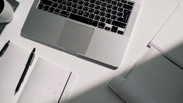 minimalist workspace featuring a laptop, a notebook, a cup of coffee, pens, and a small plant on a white desk, illuminated by natural light