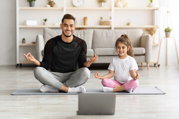 Obraz premium Relaxed arab father and little daughter having online yoga class, exercising at home together, wearing sportswear, training on yoga mat in front of laptop, enjoying workout, copy space