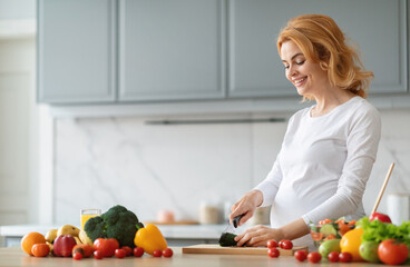 A pregnant woman smiles while chopping vegetables in her stylish kitchen. Fresh produce, including broccoli and tomatoes, is spread around her as she enjoys cooking a nutritious meal, copy space