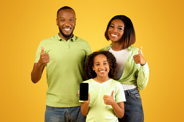 Positive african american family of three demonstrating smartphone and showing thumbs up, posing over yellow background. Happy parents and child recommending new mobile application or website