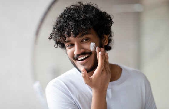 Facial skincare. Indian man applying moisturizing cream on face, looking at his reflection in bathroom mirror at home, free space. Male beauty and skin care concept