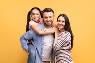 A happy family poses against a yellow background. The father holds his daughter on his back while...