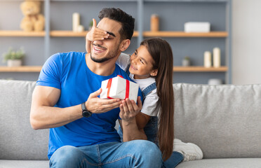 This Present Is For You. Portrait of loving girl with long hair holding and giving wrapped gift box, making surprise and covering dad's eyes, greeting with father's day or birthday, sitting on couch