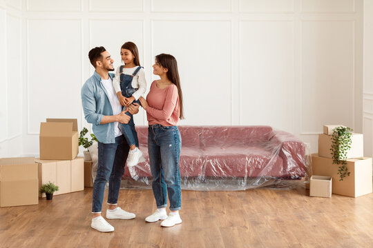House Move, Repair Concept. Full body length of cheerful young family of three people standing in new empty apartment, looking at each other, free copy space. Parents holding daughter on hands