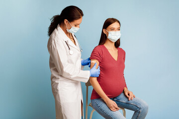 Pregnancy And Immunization Vaccination. Professional Nurse Vaccinating Pregnant Lady Sticking Plaster Strip After Vaccine Injection Against Coronavirus Over Blue Studio Background. Covid-19 Protection