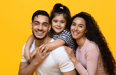 Closeup Portrait Of Cheerful Arab Family With Little Daughter Hugging And Smiling At Camera, Happy Middle Eastern Parents Bonding With Their Child While Posing Together Over Yellow Studio Background