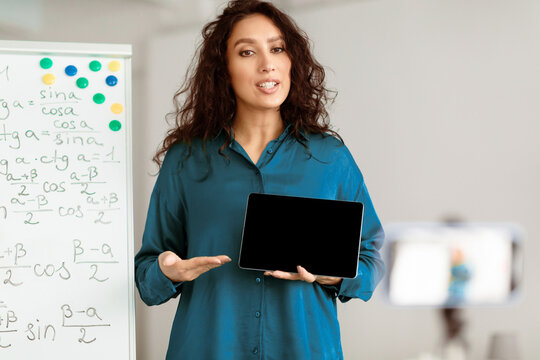 Modern Distance Learning. Lady math teacher making video conference chat with students using cellphone on tripod, holding and showing tablet with empty screen with copy space for mock up, explaining