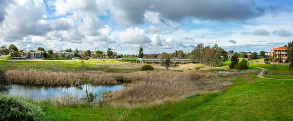 Panoramic lush wetland landscape in Tarneit with a creek, tall reeds, and a bridge connecting the green banks. Suburban houses in the neighborhood in the distance. Beautiful environment in Melbourne