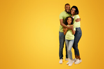 Healthy family relationship. African american dad and mom embracing smiling daughter standing over yellow studio background, crop. Full length portrait