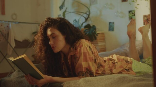 Young long-haired woman lying on stomach on bed and reading book in cozy sunlit room with plants and wall decorations, resting during day at home