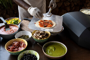 Closeup of safari camp cook making handmade pizzas with bowls of sauce and ingredients, and raw dough on pizza paddle, Lewa Conservancy, Kenya, Africa
