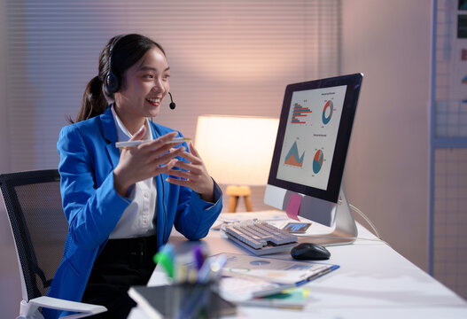 Young professional woman wearing a headset, engaging in a video call while explaining charts displayed on her computer screen during late-night hours in the office