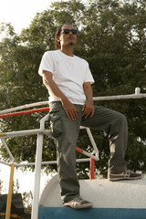 An Asian male model wearing a plain white t-shirt and black sunglasses is posing in a commercial park during the daytime. The male model's white t-shirt is being used as a mockup object for showcasing