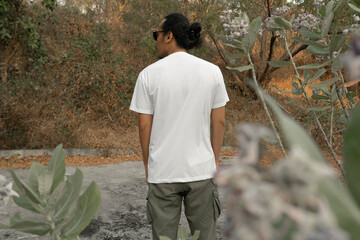 An Asian male model wearing a plain white t-shirt and black sunglasses is posing in a commercial park during the daytime. The male model's white t-shirt is being used as a mockup object for showcasing