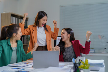 Three asian businesswomen are raising their arms and cheering in front of a laptop, celebrating a successful project or achievement in their office at night