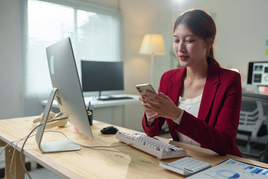 Young office worker sitting at a modern desk, using a smartphone while surrounded by a computer and business charts, embodying a productive and connected workplace atmosphere