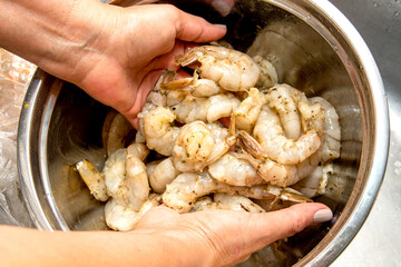 Hands holding a stainless steel container full of raw marinated shrimp. The shrimp have visible spices, ready to cook. Slightly out of focus background,