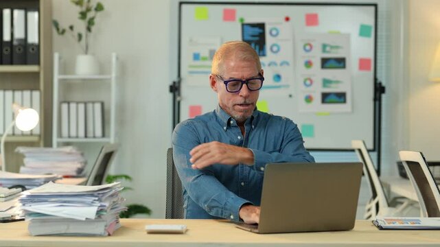 Office worker massages his shoulders and stretches his arms to relieve pain caused by long hours spent sitting at his desk working on a laptop