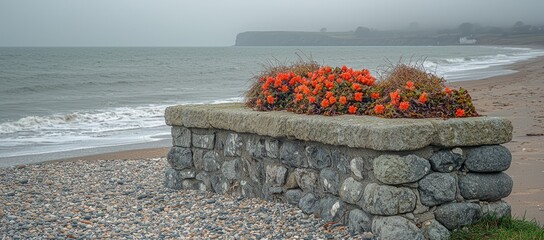 Stone wall planter with orange flowers overlooking a grey ocean beach on an overcast day.