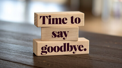 Three stacked wooden blocks display "Time to," "say," and "goodbye." in dark brown serif font on a dark wooden surface. A blurred bright interior background adds a warm, reflective tone.






