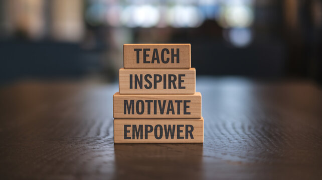 Four stacked wooden blocks display "TEACH," "INSPIRE," "MOTIVATE," and "EMPOWER" on a dark wooden surface. Sharp focus highlights the motivational words against a blurred indoor background.






