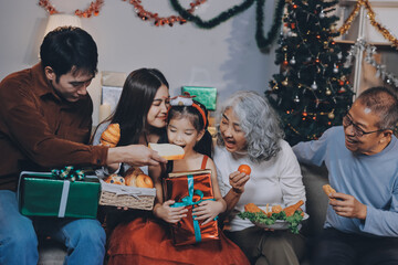 Fototapeta premium Portrait of Asian family exchanging presents during christmas at home. Attractive happy people holding gift box, celebrate holiday thanksgiving, xmas eve tradition in living room in house together.