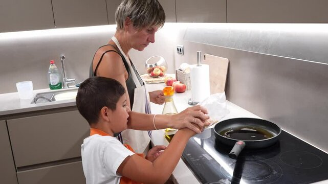 Mom carefully pouring corn kernels into a hot frying pan with oil, assisted by her young son, in a modern kitchen