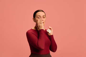 Woman in burgundy top touching eye gently with hand, holding tissue in other hand, face showing sadness or discomfort against pink background. Concept of healthcare, medicine, infection and treatment.