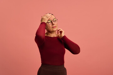Mature woman touches her forehead with hand, feeling dizzy or sick, eyes closed, against clean pink...