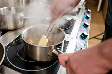 A chef's hand uses tongs to stir food in a steaming pot on a professional kitchen stove. Metal pots are visible in the background, emphasizing a busy cooking environment.