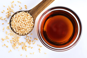Sesame oil in a glass bowl and wooden spoon with sesame seeds on white background.