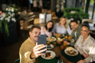 Group of friends sitting together at a restaurant, smiling and taking a selfie with their smart phone.