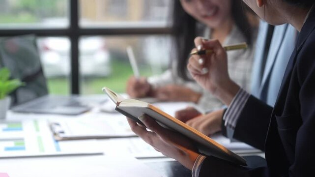 A man is writing in a notebook while two other people look on. The scene suggests a meeting or a brainstorming session where ideas are being shared and recorded