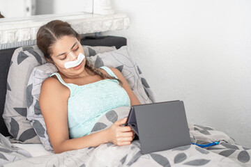 Woman resting in bed in casual clothes and bandages on her face after plastic surgery on her nose,using tablet to communicate with family, calm atmosphere and soft colors illustrate recovery process